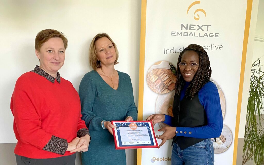 Trois femmes souriantes, vêtues de tenues professionnelles, se tiennent côte à côte en tenant un cadre lors de la cérémonie de remise du label MORE à la société Next Emballage. La scène formelle illustre la reconnaissance de l'engagement de l'entreprise pour l'innovation et le développement durable.